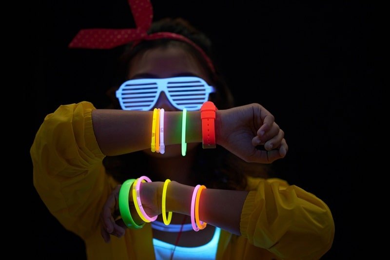 Fröhliche junge Frau mit Neonarmbändern und Brille im dunklen Raum