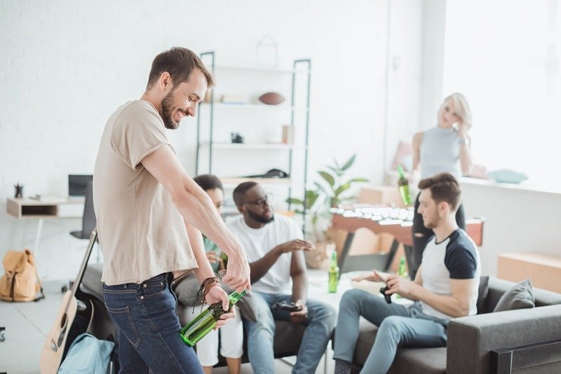 20 Partyspiele für Erwachsene Lustig, Interessant für Drinnen und Draußen junger Mann, der Bierflasche öffnet, während Freunde im Hintergrund sprechen Lustige Partyspiele für Erwachsene