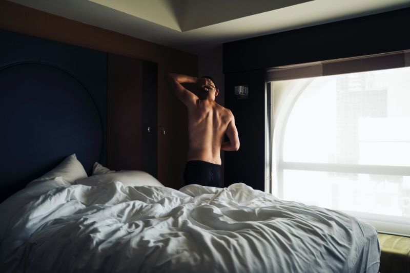 Ein toplesser Mann, der neben einem unordentlichen Bett steht