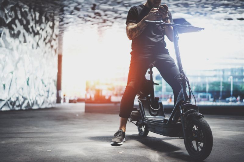 E Scooter Kinder: Alles Was Sie Wissen Sollten Bärtiger-tätowierter-männlicher-Hipster-mit-Handy-nach-dem-Fahren-mit-dem-Elektroroller-in-der-Stadt-min