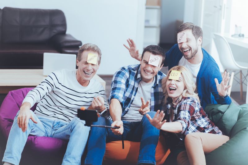 Fröhliche-lächelnde-Kollegen-die-Aufkleber-auf-der-Stirn-haben-und-Selfies-machen-während-sie-einen-Spieleabend-haben