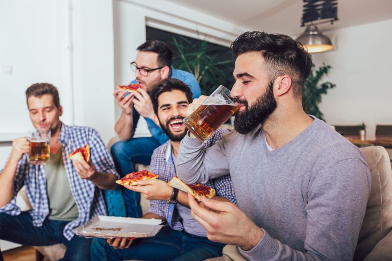 Spieleabend: Organisieren Sie Das Perfektes Spieltreffen Gruppe-von-vier-männlichen-Freunden-die-Bier-trinken-und-zu-Hause-Pizza-essen