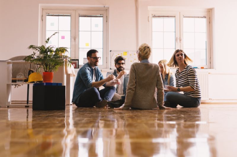 Moderne fröhliche junge Leute die zusammen auf dem Büroboden arbeiten und Zusammengehörigkeit erhöhen
