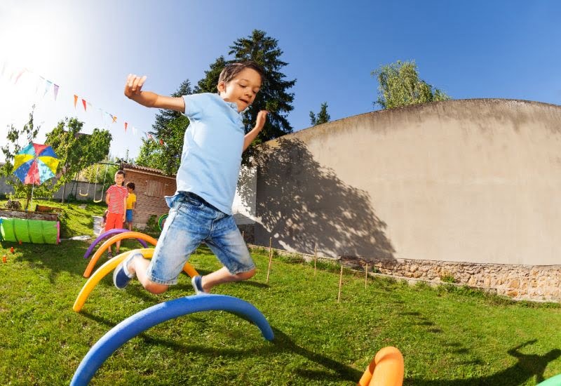 Netter-hübscher-Junge-im-blauen-T-Shirt-in-einer-Gruppe-von-Kinderfreunden-springt-über-Barriere-in-dem-Hindernisparcours
