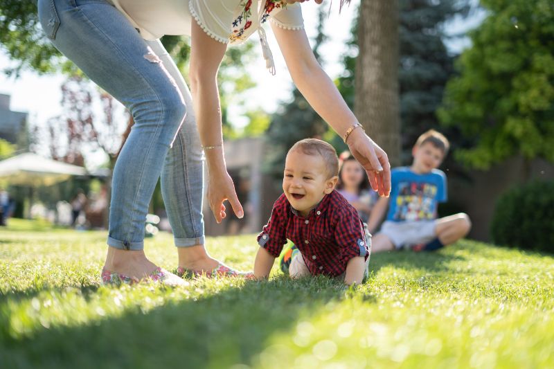 Tagesmutter: Beratung, Qualifizierung Und Infos Für Die Eltern Frau-in-der-weißen-Bluse-und-in-den-blauen-Jeans-die-einem-Baby-helft-auf-grünem-Gras-zu-kriechen