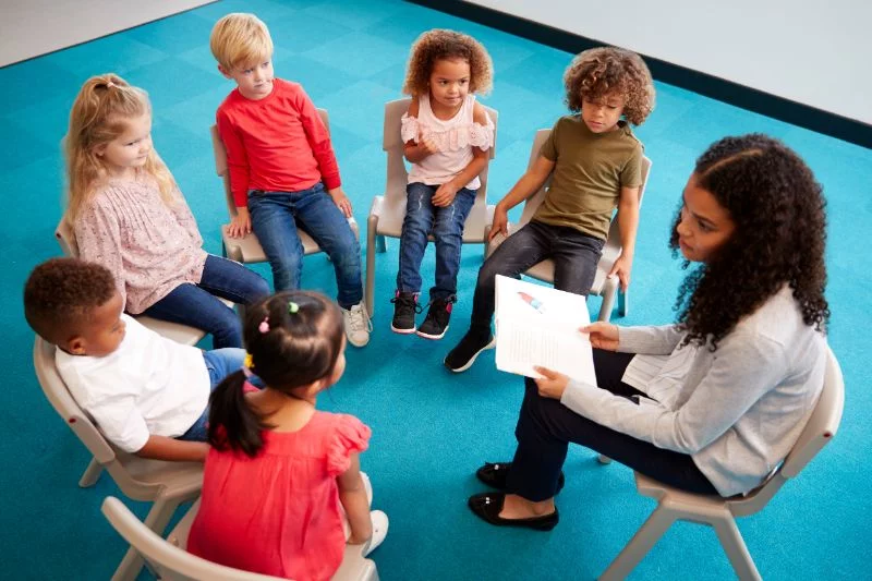 Junge-weibliche-Schullehrerin-die-ein-Buch-zu-den-kleinen-Schulkindern-liest-und-auf-Stuhlen-in-einem-Kreis-im-Klassenzimmer-sitzt-und-zuhort