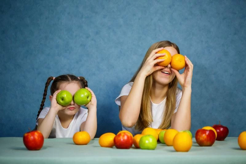 Kleines-Madchen-und-Mutter-spielen-mit-Fruchten.-Sie-tragen-weise-T-Shirts.