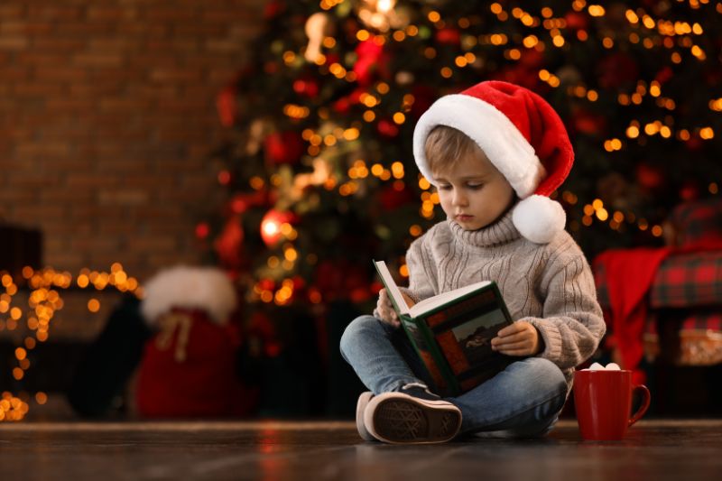 Kleiner-Junge-in-der-Weihnachtsmannmutze-die-Buch-nahe-Weihnachtsbaum-liest
