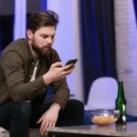 Mann mit Bart hält ein Handy und trinkt Bier
