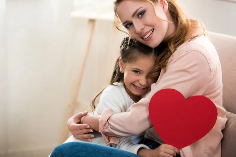 Gluckliche-Mutter-und-Tochter-mit-rotem-Herzsymbol-das-Kamera-umarmt-und-lachelt