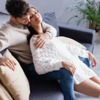 Junger Mann, der lächelnde Frau im Pullover auf Sofa mit Kissen umarmt