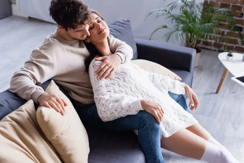 Junger Mann, der lächelnde Frau im Pullover auf Sofa mit Kissen umarmt