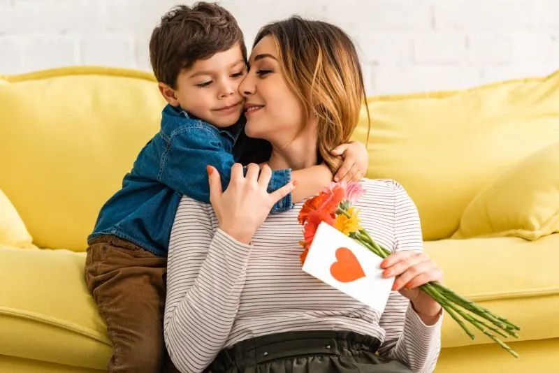Netter Junge, der Mutter umarmt, die Blumen und Muttertagskarte mit Herzsymbol hält