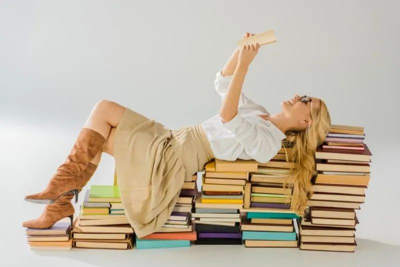 Schöne blonde Frau in den Gläsern, die auf Stapel Bücher lesen und legen