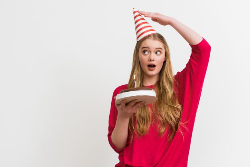 Überraschtes Mädchen, das Partykappe berührt und Geburtstagstorte hält