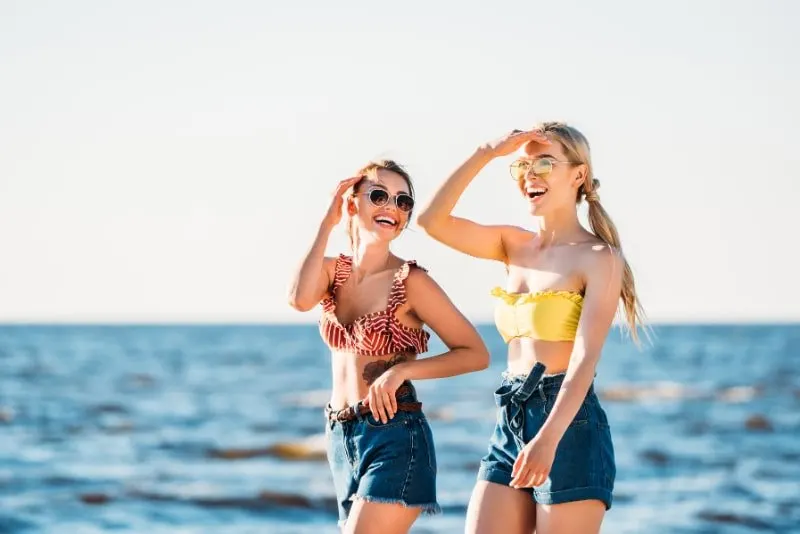 Glückliche junge Frauen in der Sonnenbrille, die zusammen am Strand gehen
