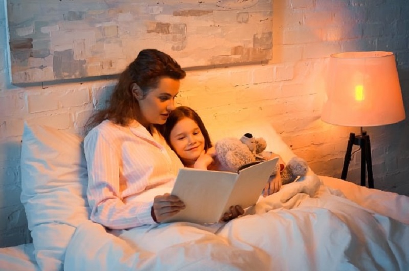 Glückliches Mädchen mit Teddybär liegend nahe Mutter, die Buch auf Bett liest