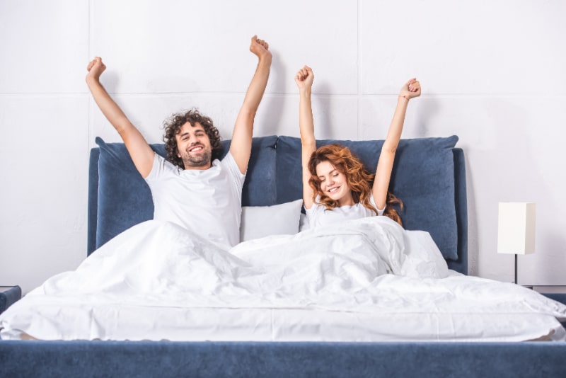Gluckliches-junges-Paar-das-Arme-streckt-und-zusammen-im-Schlafzimmer-aufwacht