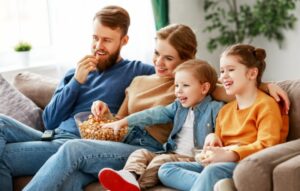 Glückliche junge Eltern mit süßen Kindern, die auf der Couch im Wohnzimmer sitzen und Fernsehen, Film, Netflix schauen