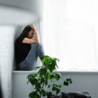 depressive junge Frau weint, während sie auf der Fensterbank sitzt und die Hände am Kopf hält