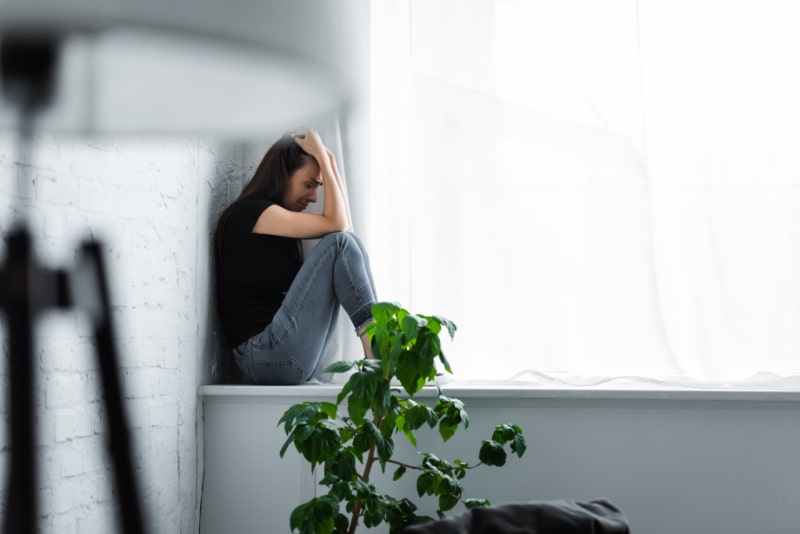 depressive junge Frau weint, während sie auf der Fensterbank sitzt und die Hände am Kopf hält