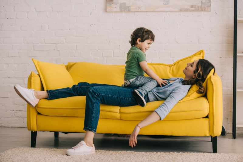 Kleiner Junge sitzt auf müder Mutter, während sie auf einem gelben Sofa im Wohnzimmer liegt