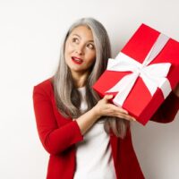 Elegante asiatische Seniorin, die Geschenkbox rüttelt und versucht zu erraten, was im Inneren vorhanden ist und über weißem Hintergrund steht. Feiertage und Feierkonzept
