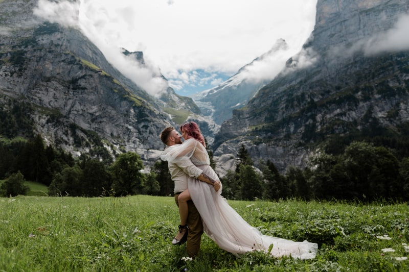 Gluckliche-Braut-im-Hochzeitskleid-und-Brautigam-die-Berge-und-Wolken-in-den-Alpen-umarmen