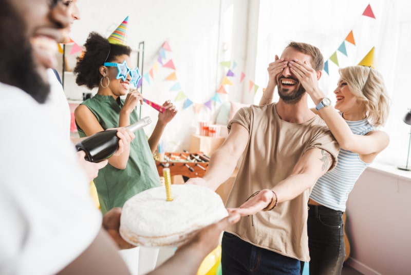 Lachelnde-junge-Leute-die-die-Augen-eines-jungen-Freundes-bedecken-und-ihn-mit-Geburtstagskuchen-begrusen