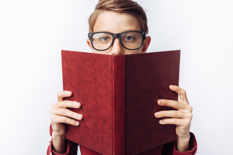 Portrat-eines-positiven-und-emotionalen-Schuljungen-der-hinter-einem-Buch-weisem-Hintergrund-Brille-rotem-Hemd-Geschaftsthema-hervorschaut