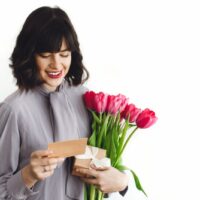 Schöne brünette Mädchen mit Blumenstrauß aus Tulpen, Geschenkbox und Grußkarte auf weißem Hintergrund drinnen, Platz für Text. Glückliche junge Frau mit Blumen. Schönen Muttertag.