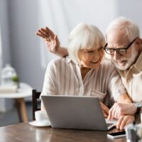 Selektiver Fokus eines positiven Seniorenpaares mit Videoanruf auf Laptop in der Nähe von Smartphone und Kaffee auf dem Tisch