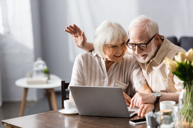 Selektiver Fokus eines positiven Seniorenpaares mit Videoanruf auf Laptop in der Nähe von Smartphone und Kaffee auf dem Tisch