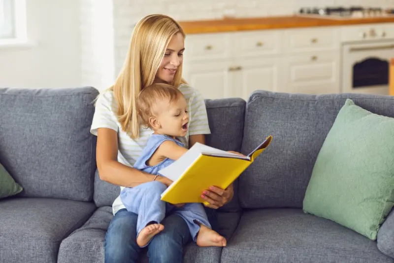 Junge-Mama-liest-ihrem-kleinen-Kind-ein-Marchenbuch-vor-das-zu-Hause-auf-der-Couch-sitzt