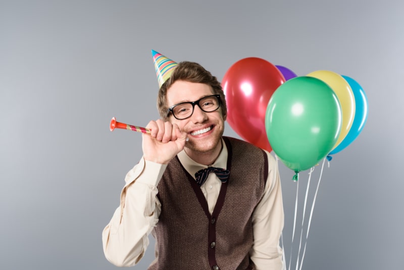 Lachelnder-Mann-mit-Brille-und-Partymutze-mit-bunten-Luftballons-und-Partyhorn-auf-grauem-Hintergrund
