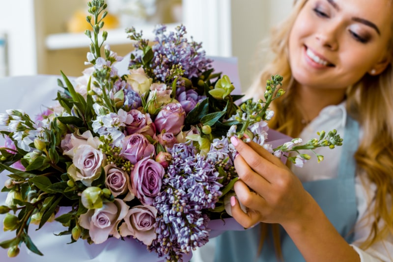 Lachelnde-Floristin-arrangiert-Blumenstraus-mit-Flieder-und-Rosen-im-Blumenladen